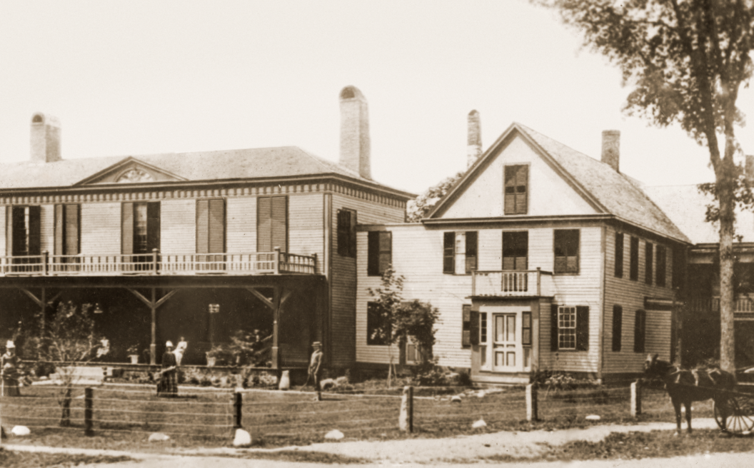 Sepia photo of a large, historic wooden building with porches, people standing outside, and a horse-drawn carriage in front.