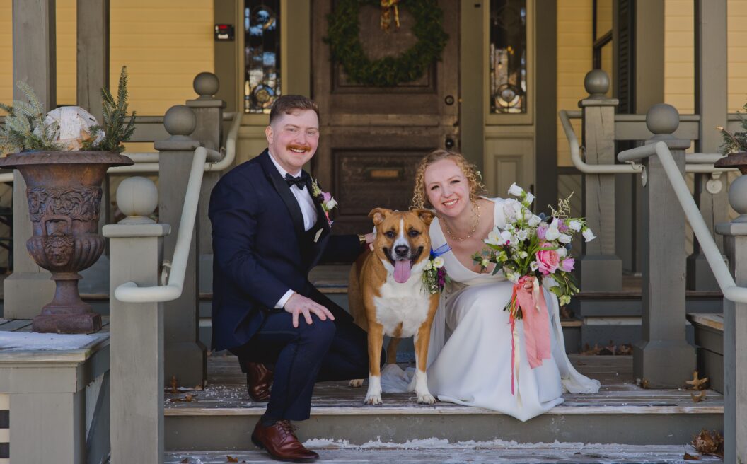 A bride and groom in formal wear pose smiling with a brown and white dog on the snowy steps of a decorated porch.