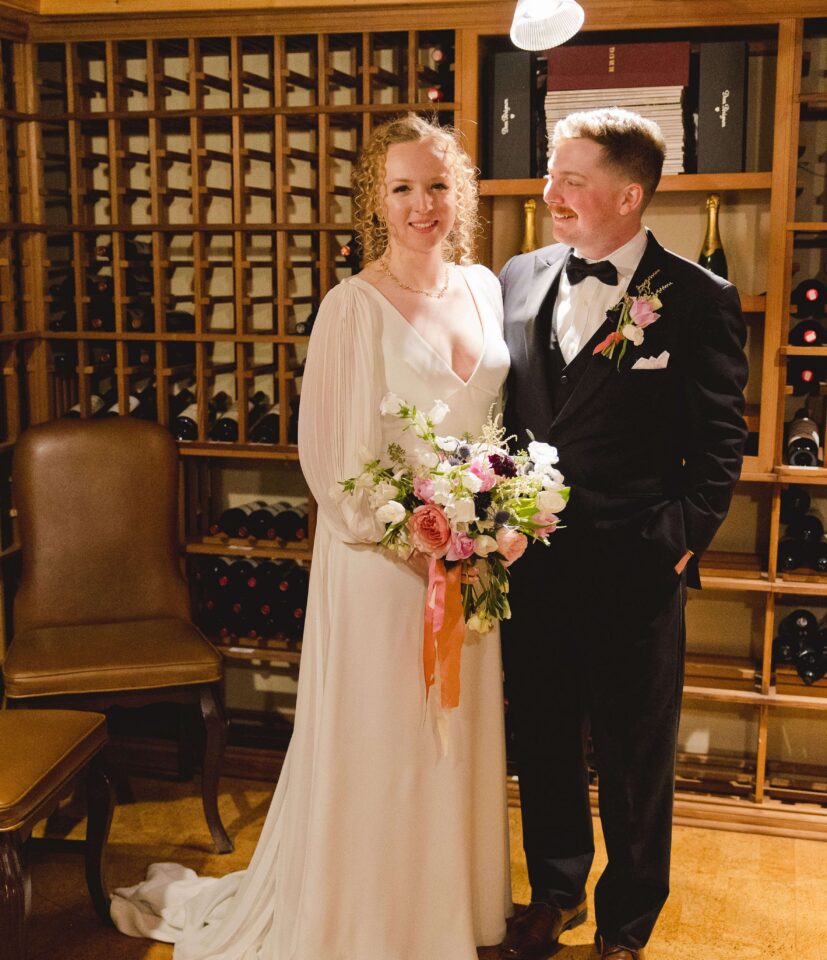 A bride in a white dress holds a bouquet and stands next to a groom in a tuxedo in a wine cellar, both smiling.