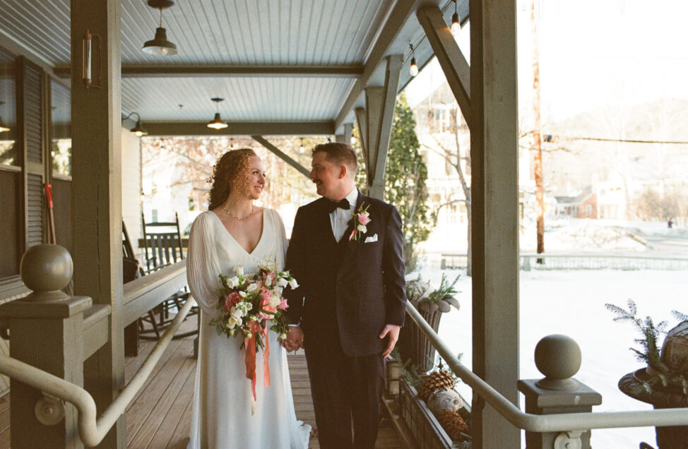 A bride and groom hold hands and smile at each other while standing on a porch decorated with flowers.