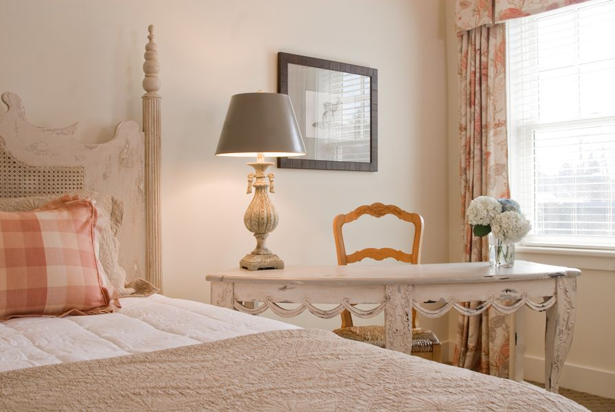 A cozy bedroom with a carved bed, plaid pillow, white desk, lamp, wooden chair, flowers in a vase, and a window with floral curtains.
