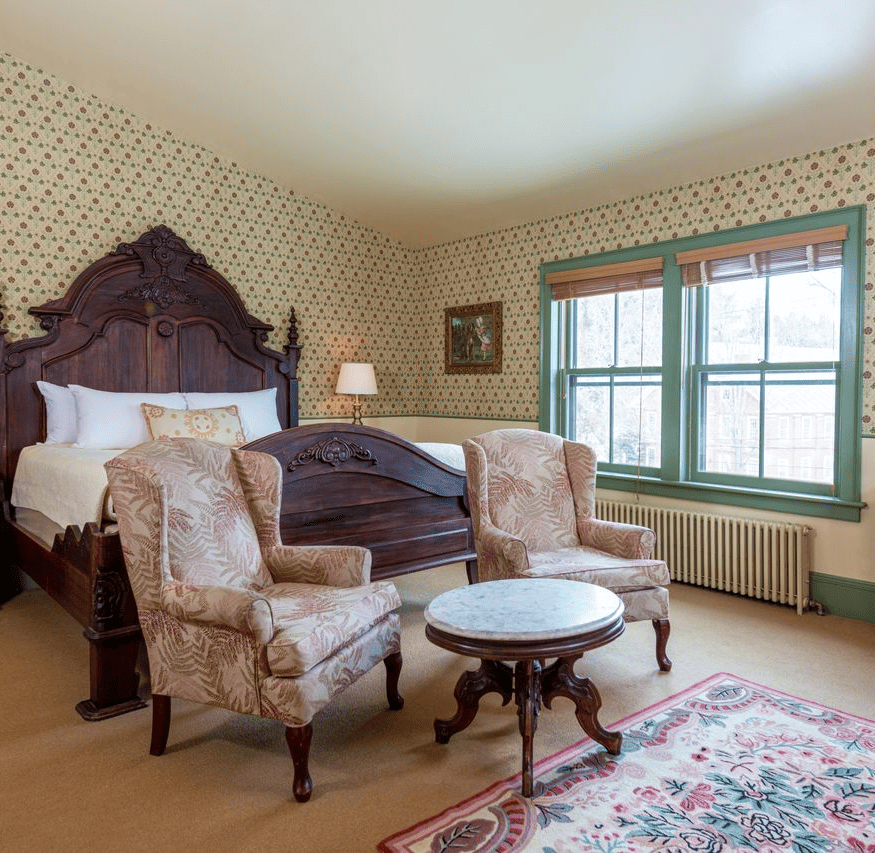 Victorian-style bedroom with ornate wooden bed, two armchairs, marble table, dresser, floral wallpaper, and large windows letting in natural light.