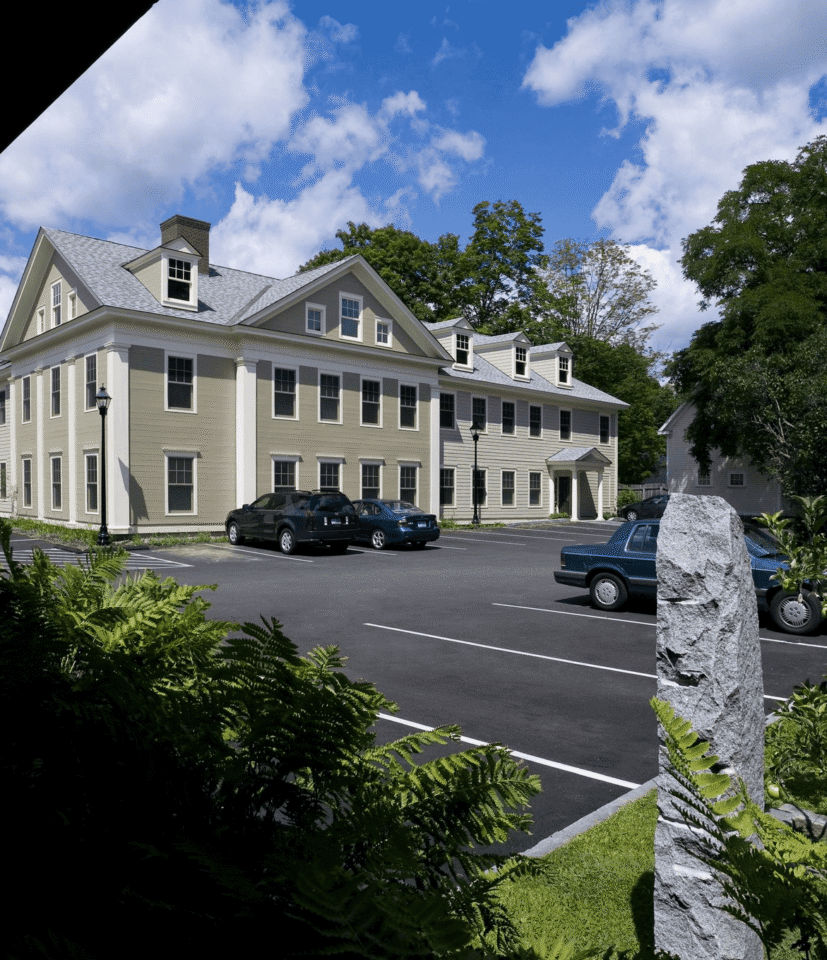 A beige, two-story building with dormer windows sits by a parking lot with cars, surrounded by greenery under a partly cloudy blue sky.