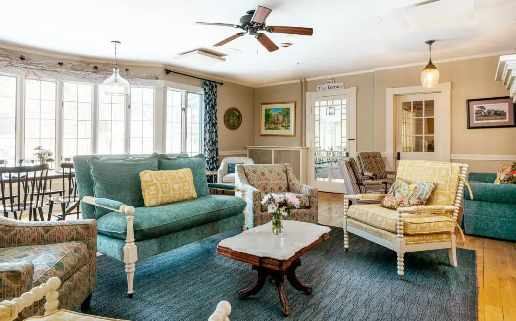 Bright living room with vintage-style sofas, armchairs, a marble coffee table, large windows, wooden floor, and ceiling fan. Flowers decorate the tables.