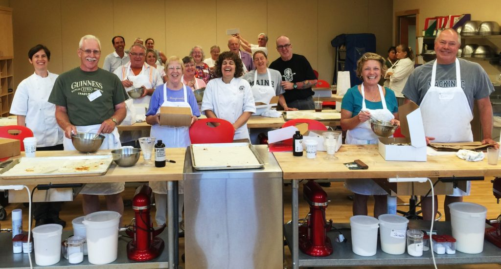 A group of adults in a kitchen classroom smile at the camera, standing behind tables with baking supplies and mixing bowls.