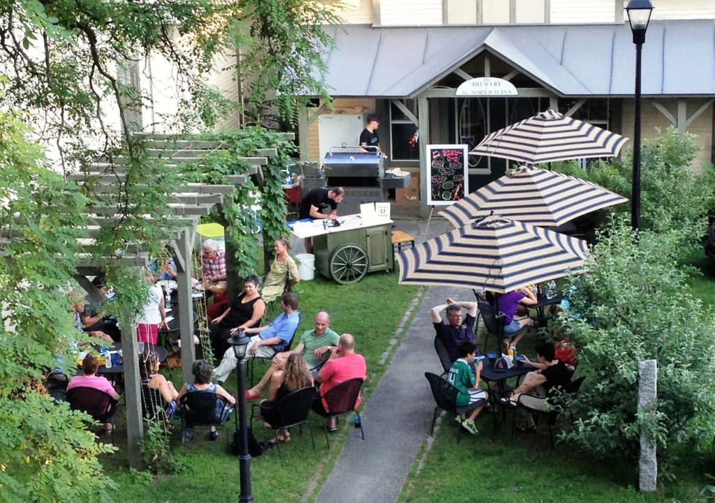 People sit at outdoor tables under striped umbrellas, socializing and eating near a food cart in a garden setting beside a building.