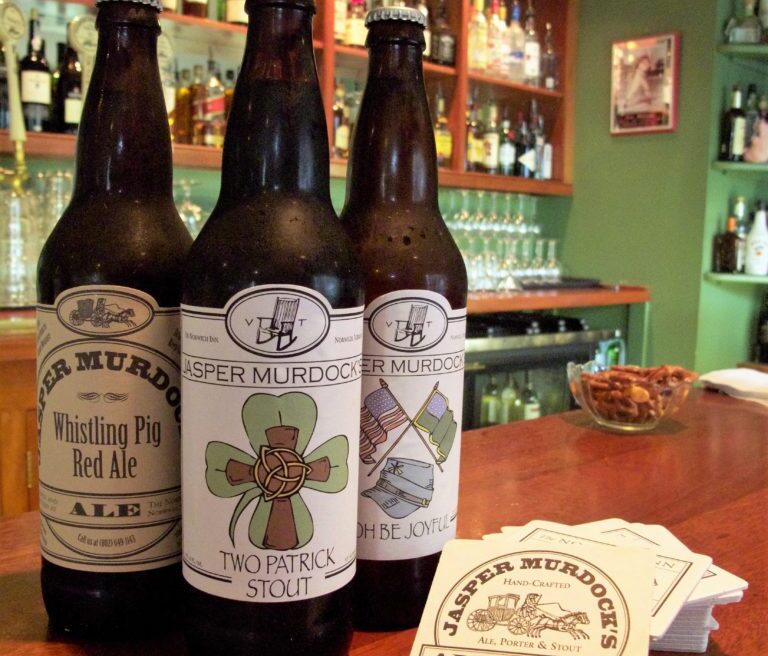 Three Jasper Murdocks Alehouse beer bottles on a bar, with coasters and a bowl of snacks nearby. Shelves of glasses and bottles in the background.