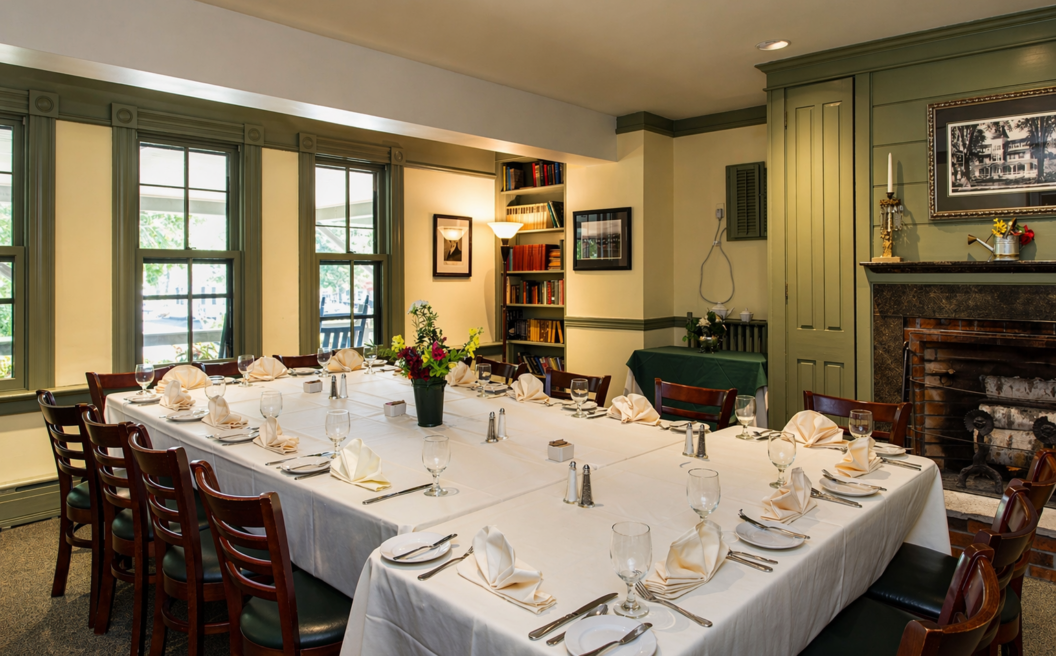 Elegant dining room with a U-shaped table set for a meal, decorated with flowers, surrounded by wooden chairs, bookshelves, and a fireplace.
