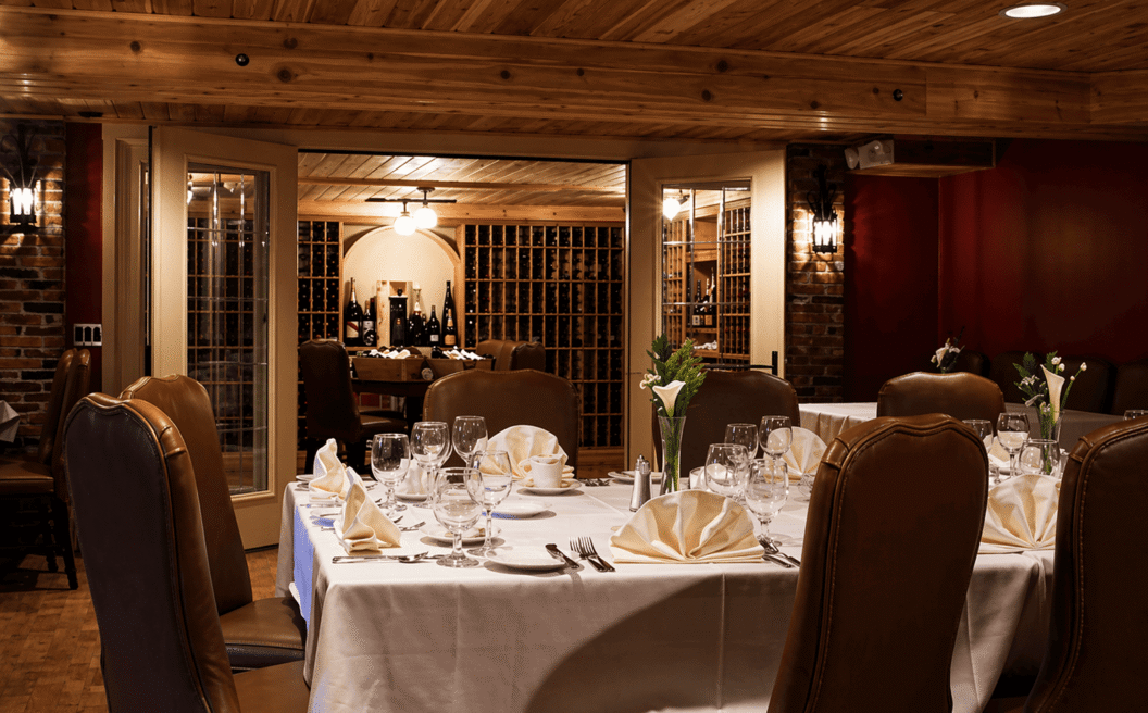 Elegant dining room with set tables, leather chairs, white linens, and a view into a wine cellar with bottles displayed on wooden shelves.