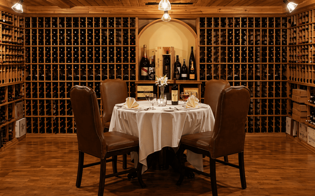 Round table with four leather chairs, set for dinner, in an elegant wine cellar lined with shelves of wine bottles and wooden boxes.