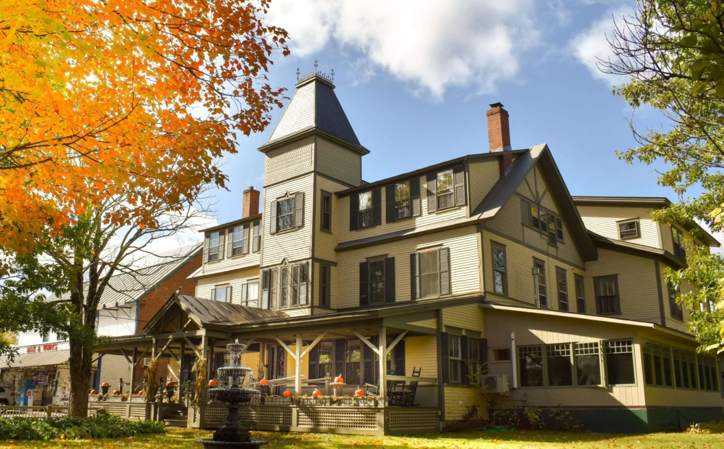 A large Victorian house with a porch and tower, surrounded by autumn trees and fallen leaves, under a partly cloudy sky.