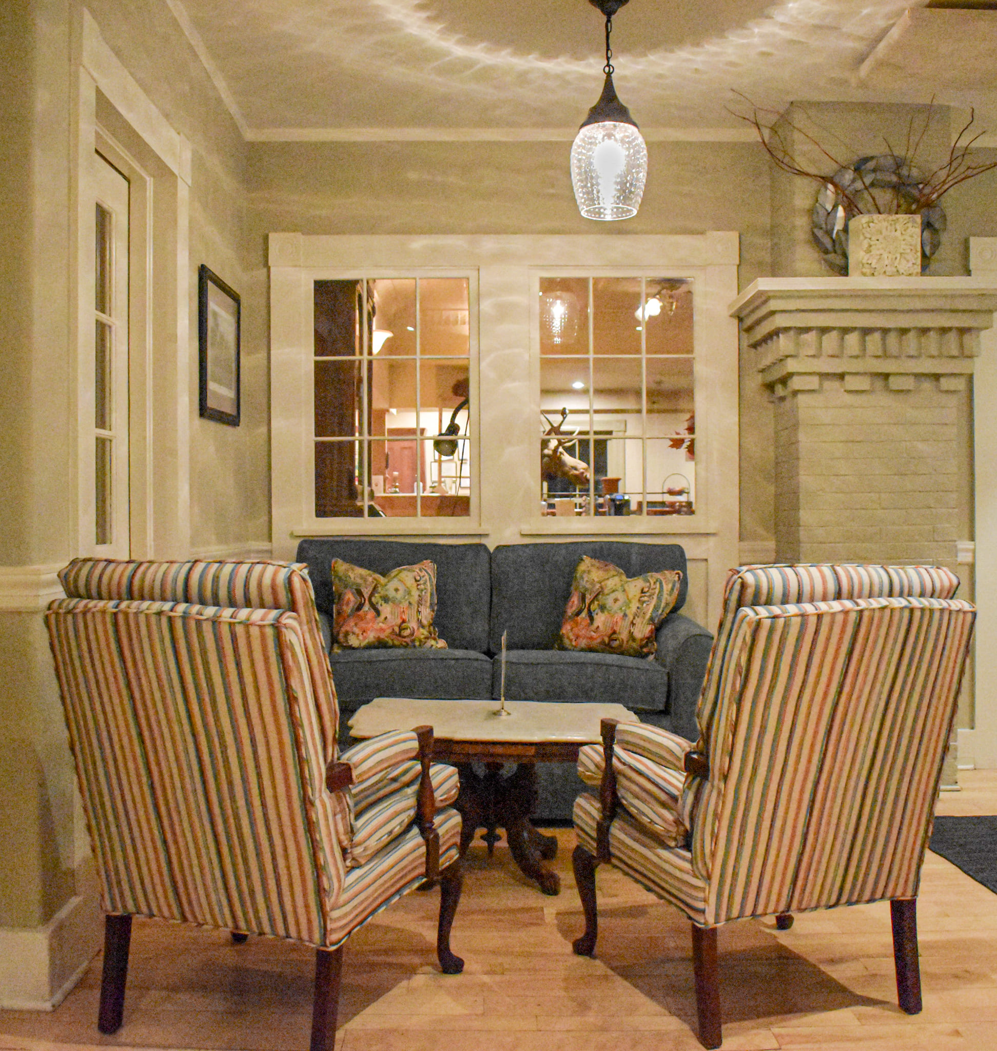 Cozy living room with a blue sofa, striped armchairs, a small table, fireplace, and a hanging light. Mirrors reflect a warmly lit interior.