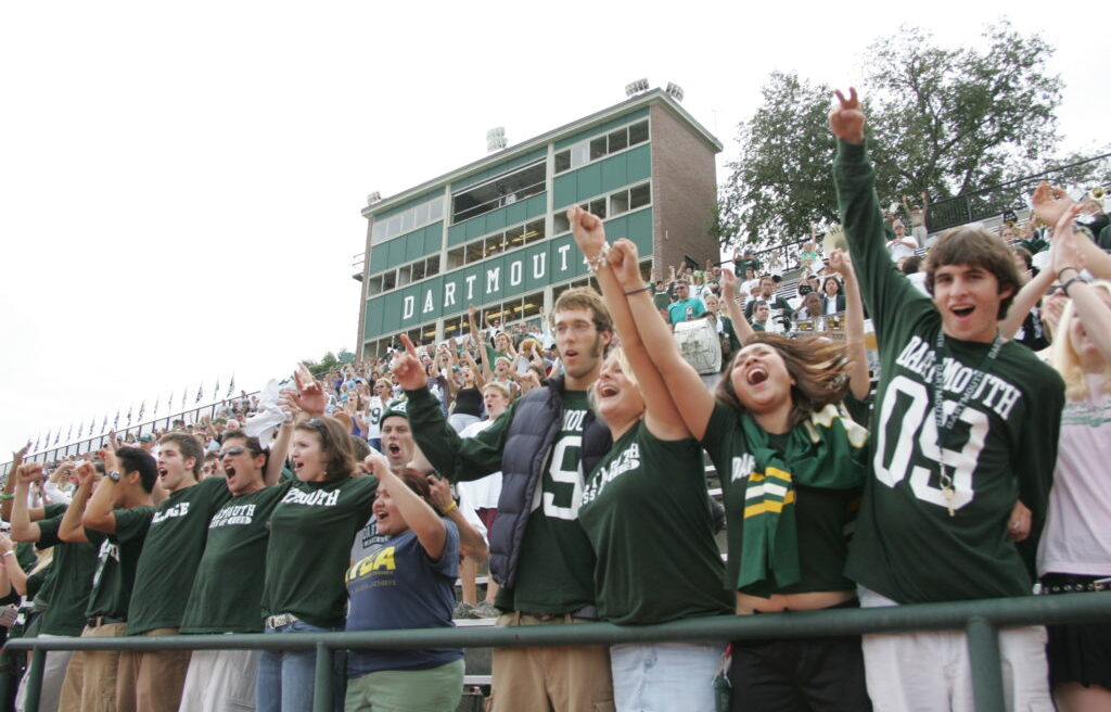A cheering crowd of young people in Dartmouth shirts stands in front of a stadium, raising their arms and smiling excitedly.