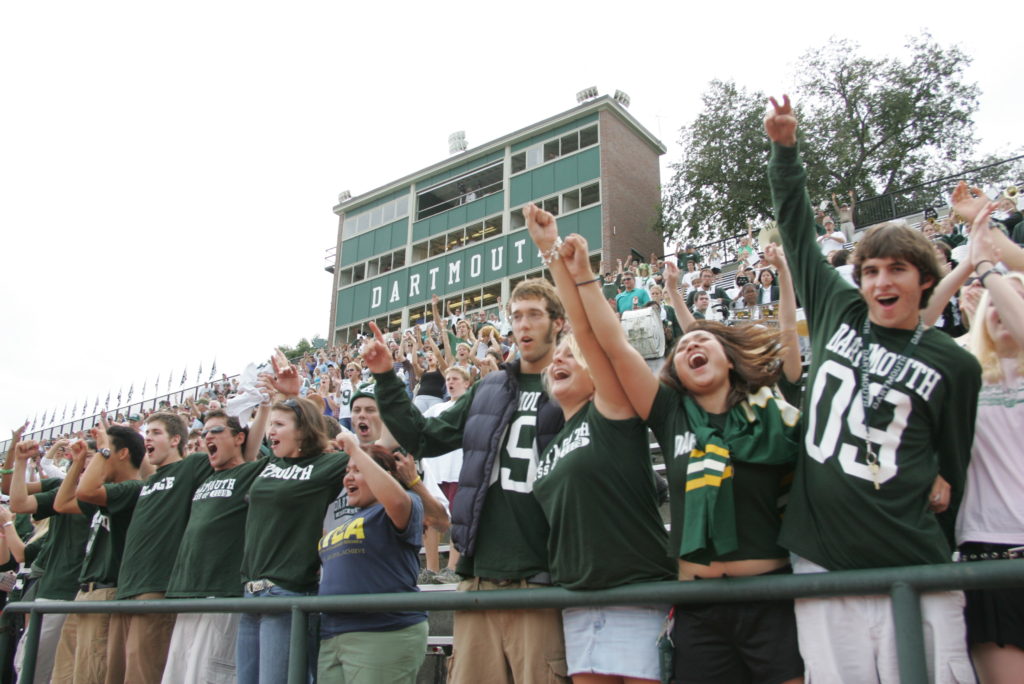 A cheering crowd of young people in Dartmouth shirts stands in front of a stadium, raising their arms and smiling excitedly.