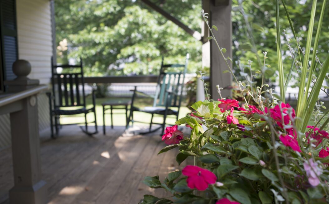 Pink flowers in focus on a wooden porch, with two rocking chairs and green trees blurred in the background.