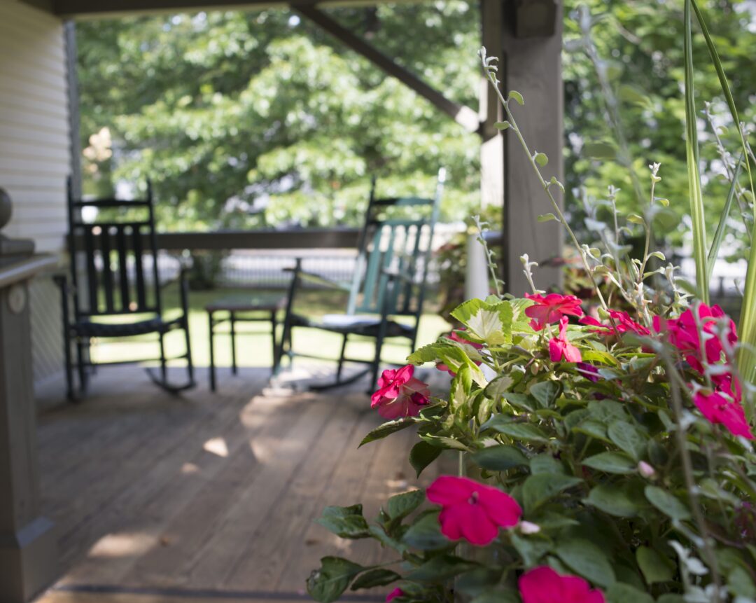 Pink flowers in focus on a wooden porch, with two rocking chairs and green trees blurred in the background.