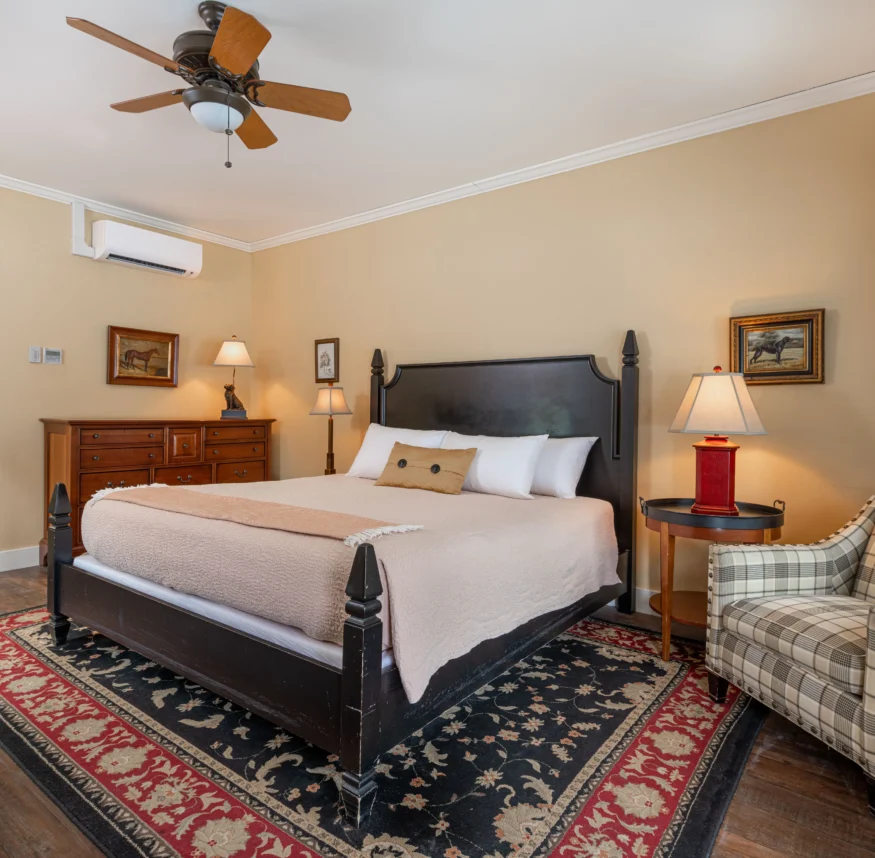 A cozy bedroom with a large bed, plaid armchair, wooden dresser, lamps, ceiling fan, and an attached bathroom visible through an open door.