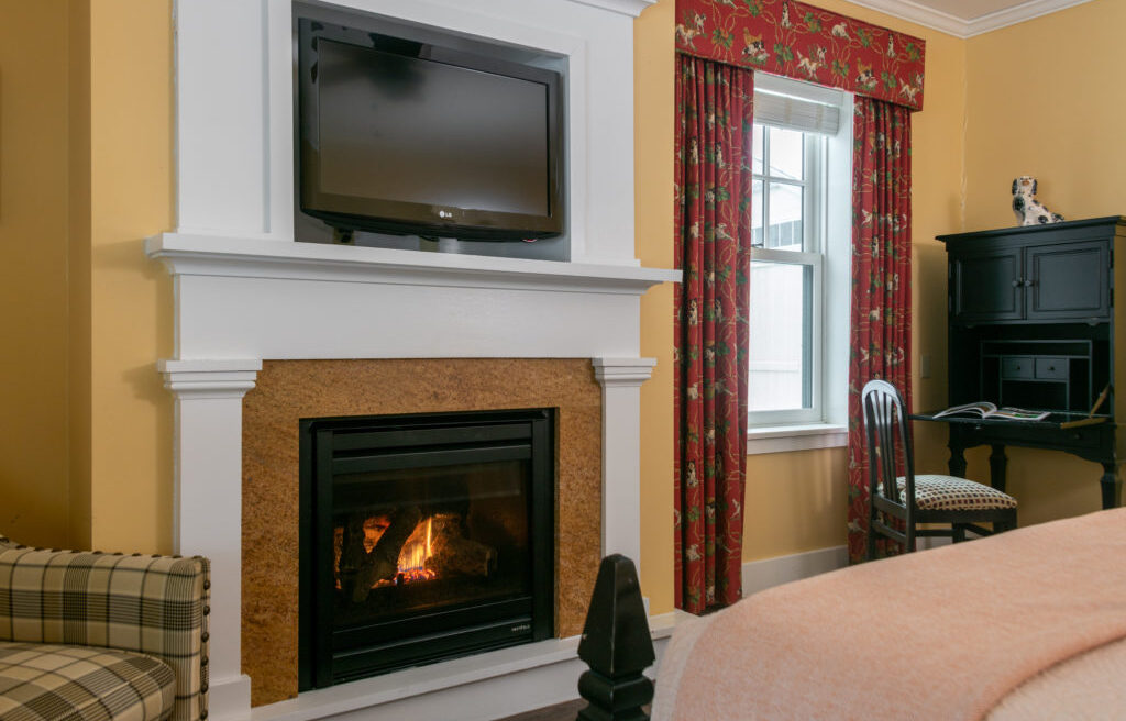 A cozy room with a fireplace, wall-mounted TV, red curtains, window, black desk, chair, plaid armchair, and part of a bed with a peach blanket.