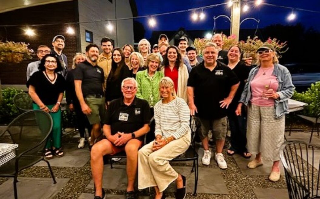 A group of people smile and pose together outdoors at night under string lights, with two people seated at the front and others standing behind them.