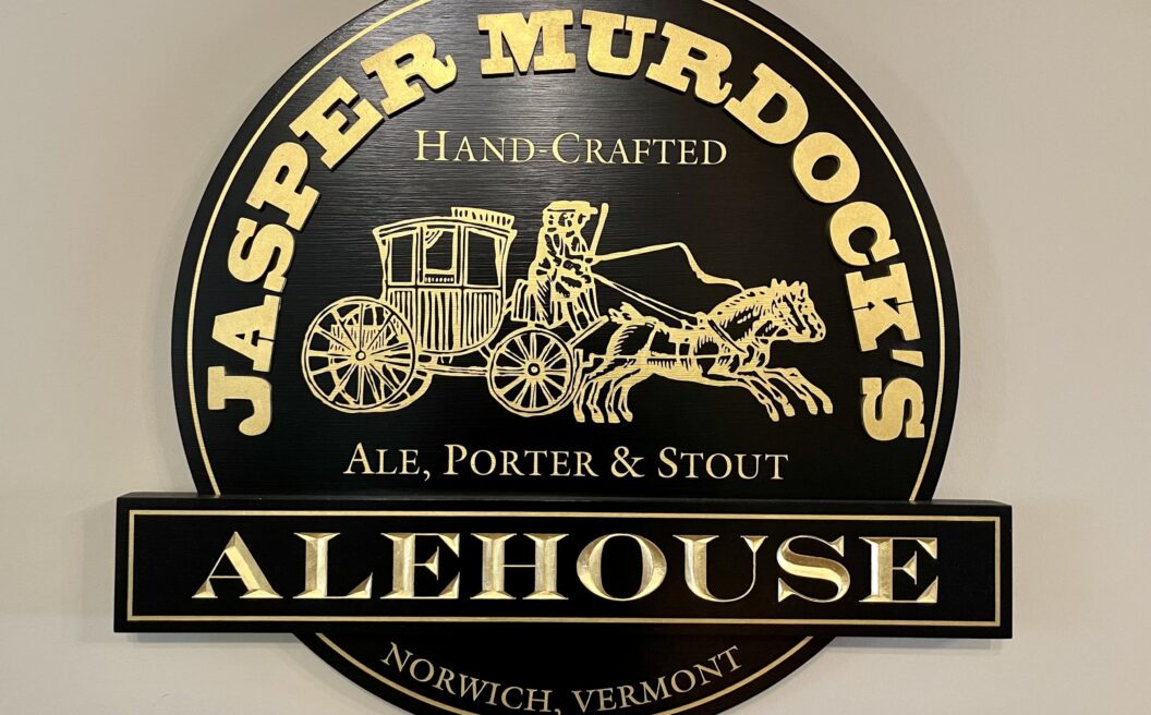 Round black and gold sign for Jasper Murdocks Alehouse, featuring a horse-drawn carriage illustration and text about ale, porter, and stout.