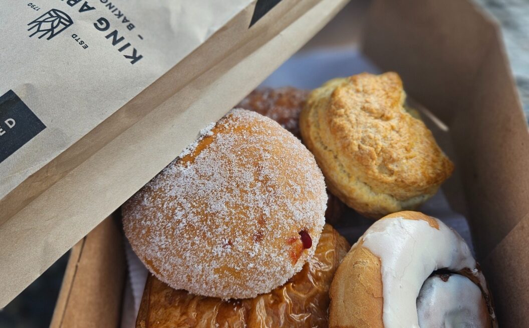 A box of assorted pastries, including a sugared donut, cinnamon roll, biscuit, and croissant, partially covered by a bakery paper bag.