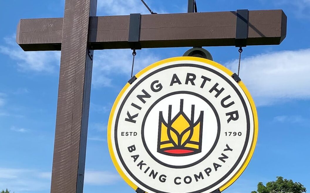 King Arthur Baking Company sign with logo, listing bakery, café, school, and store, against a blue sky and green trees.