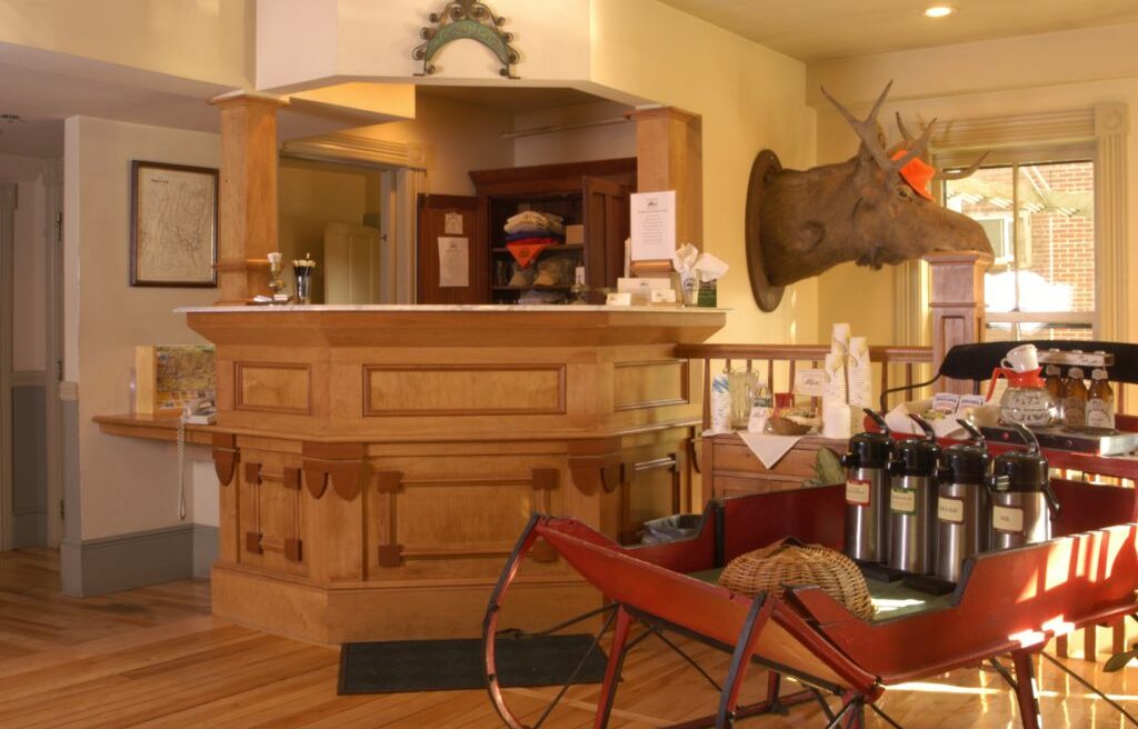 A cozy lodge lobby with a wooden reception desk, mounted moose head, coffee dispensers, and a red sleigh on a hardwood floor.