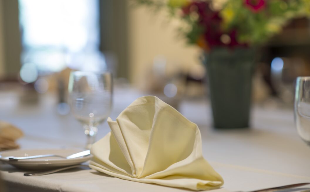 A folded cream napkin on a set dining table with glassware and a blurred vase of flowers in the background.