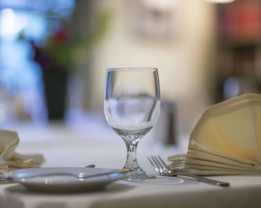 A clear wine glass, utensils, and a folded napkin are arranged on a white tablecloth in a softly lit restaurant setting.
