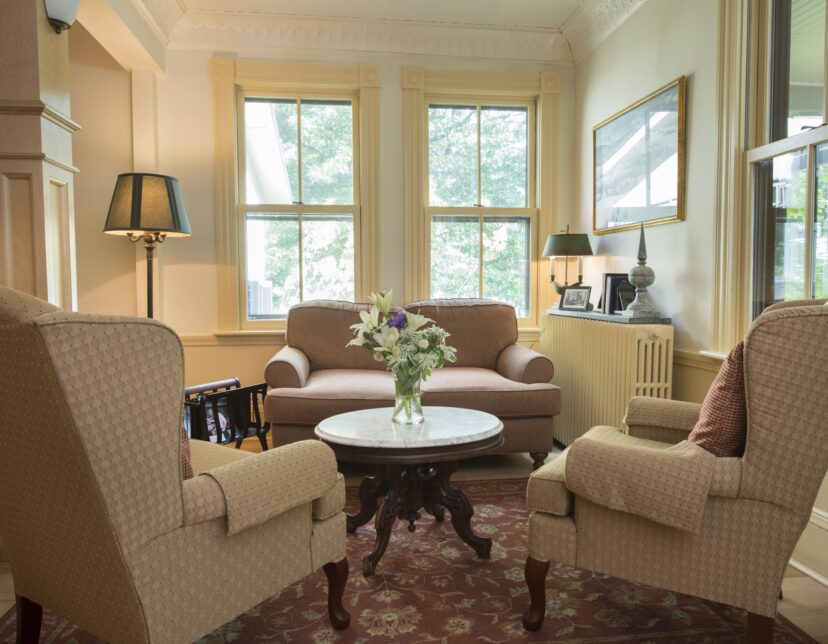 A cozy living room with beige armchairs, a sofa, a round table with flowers, large windows, and soft natural light.