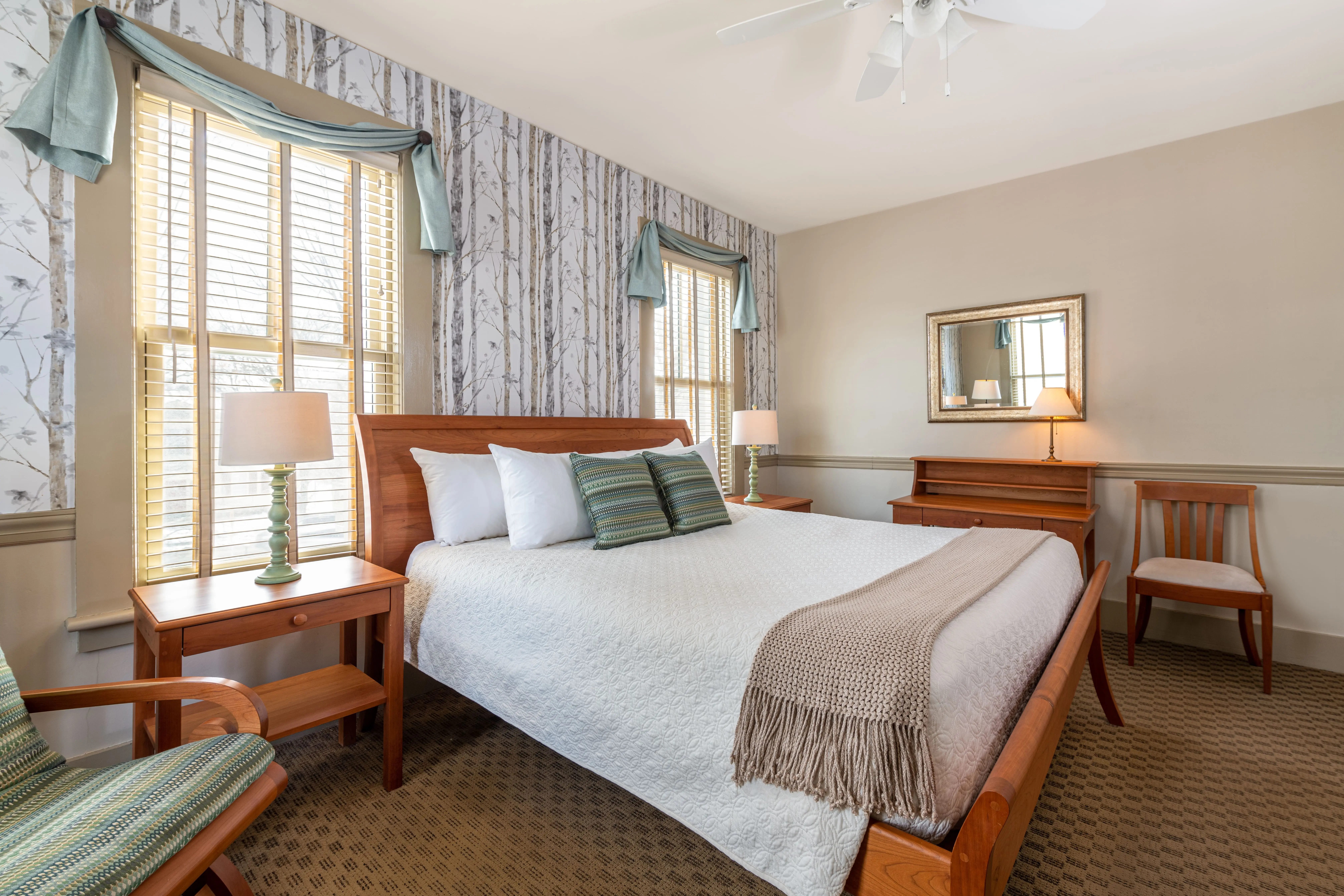 A cozy bedroom with a large bed, wooden furniture, two lamps, a mirror, and windows with patterned curtains and natural light.