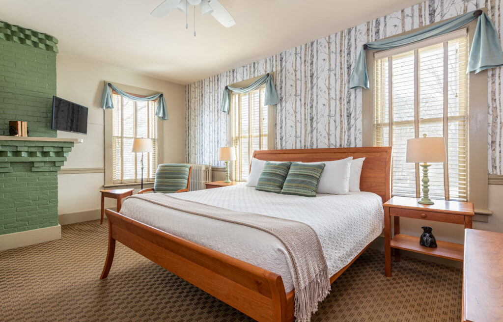 Bright bedroom with a wooden bed, green-accented pillows, lamps, a green fireplace, and birch tree wallpaper, with sunlight coming through windows.