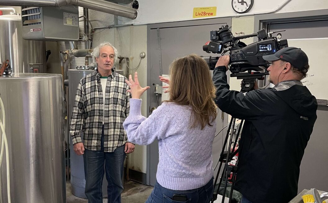 A woman interviews a man in a plaid shirt while a cameraman films in an industrial kitchen with metal tanks, white buckets, and visible pipes.