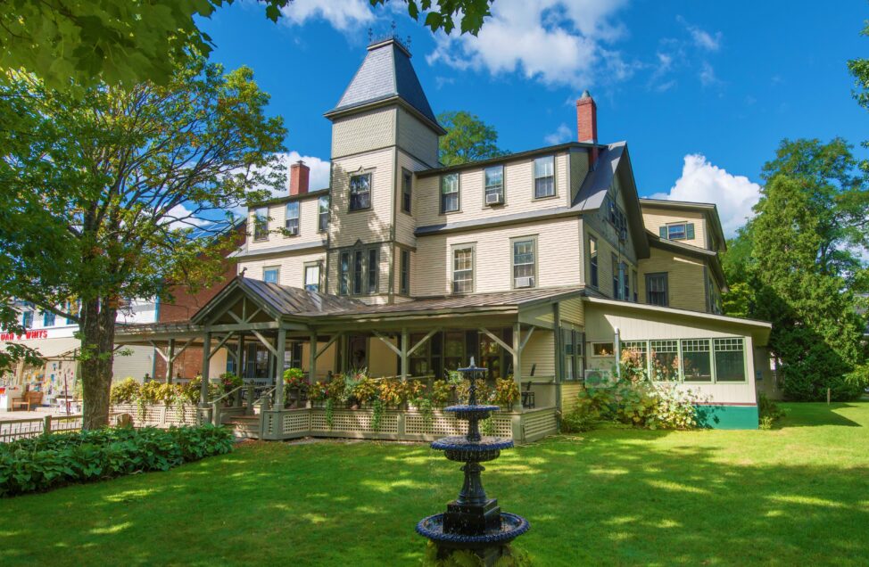 A large Victorian-style house with a covered porch, surrounded by green lawn and trees, featuring a black tiered fountain in the foreground.