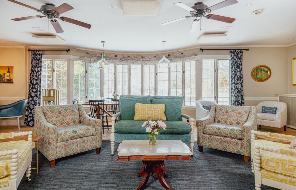 Bright living room with patterned armchairs, green sofa, marble coffee table with flowers, large windows, ceiling fans, and blue curtains.