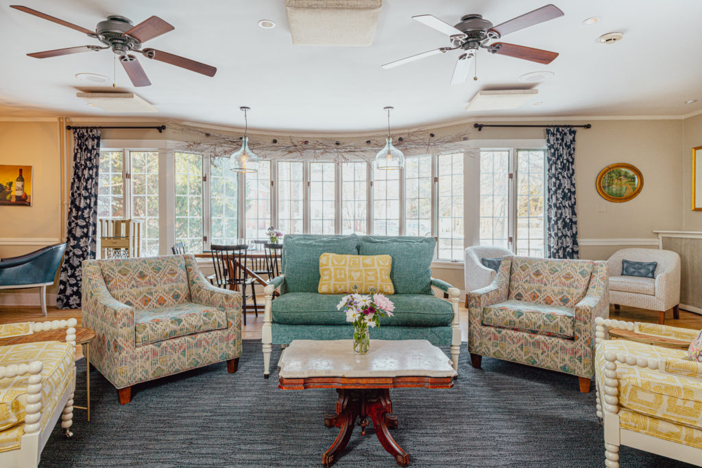 Bright living room with patterned armchairs, green sofa, marble coffee table with flowers, large windows, ceiling fans, and blue curtains.