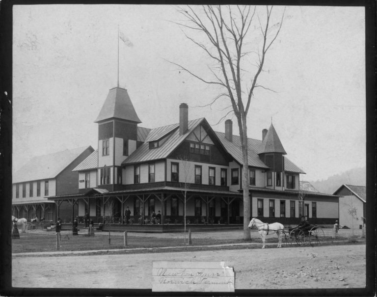 Black-and-white photo of a large Victorian-style building with a wraparound porch, people outside, and a horse-drawn carriage in front.