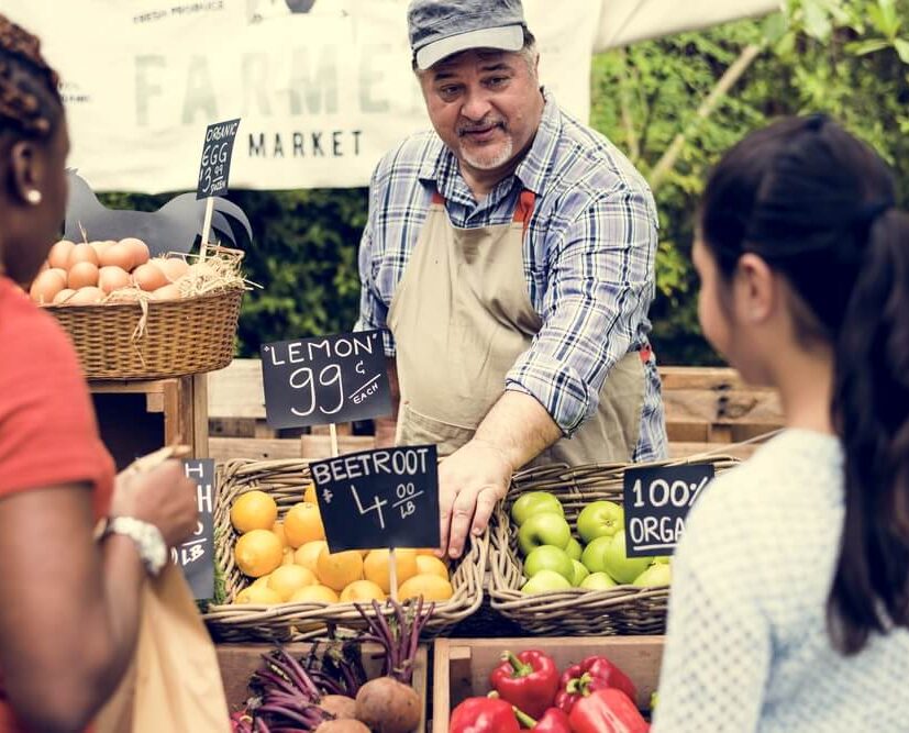 A vendor in an apron sells fresh produce to two women at a farmers market, with fruits and vegetables displayed on the table.