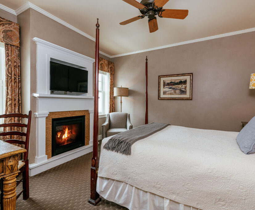 Cozy bedroom with a four-poster bed, fireplace, TV, desk, chair, and two windows with drapes. Warm lighting and neutral colors create a relaxing space.