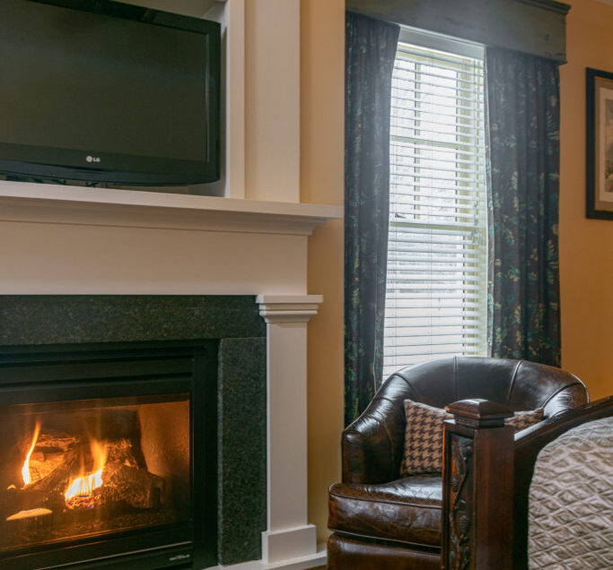 A cozy room with a lit fireplace, a leather armchair, a bed, a window with curtains, and a TV mounted above the fireplace.