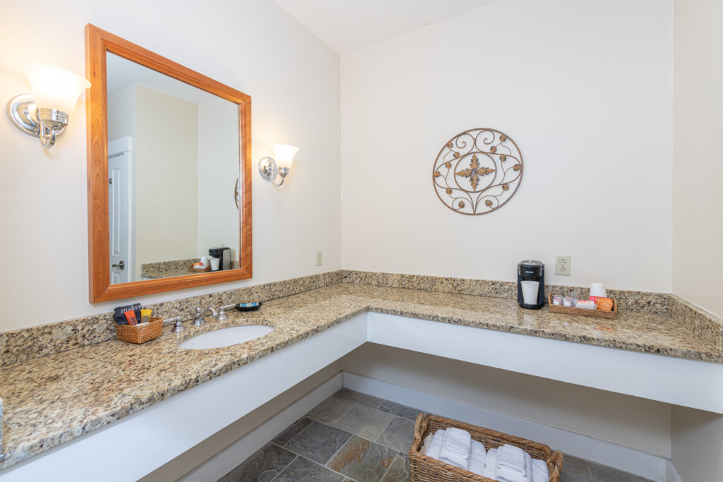 Bathroom with a granite countertop, sink, mirror, wall art, coffee maker, toiletries, and a basket with towels on a tiled floor.