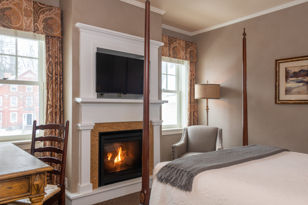 Cozy bedroom with a fireplace, TV above mantle, armchair, wooden desk, and bed with gray blanket; windows let in natural light.