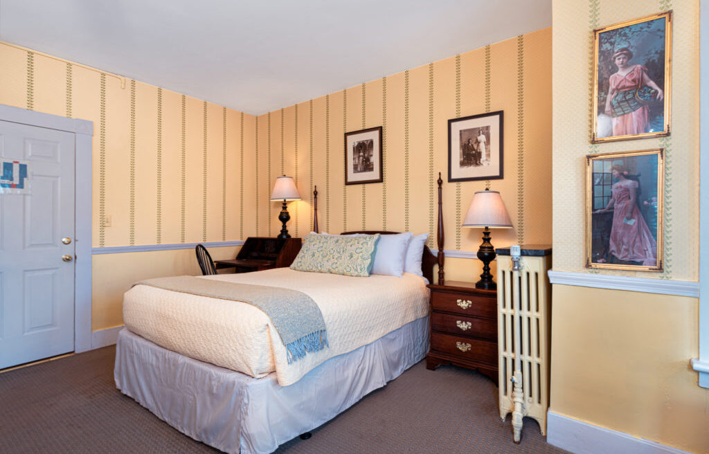 Cozy bedroom with striped yellow wallpaper, a bed, two nightstands with lamps, framed photos, and a small desk and chair in the corner.