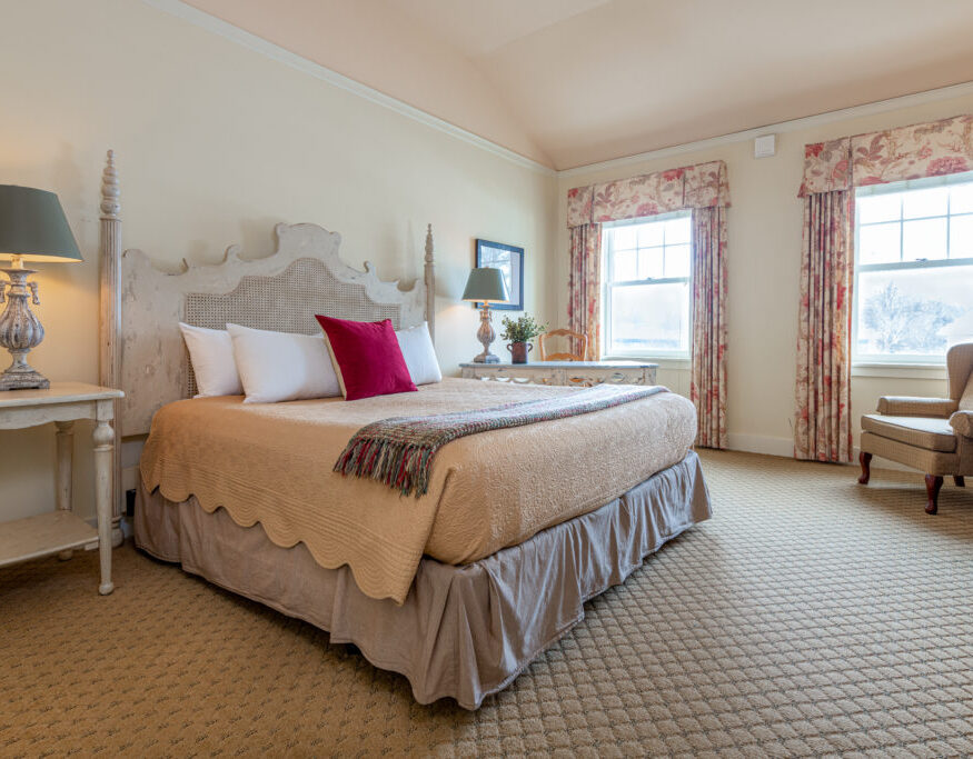A cozy bedroom with a large bed, beige bedding, red accent pillow, two windows with floral curtains, lamps, and an armchair in the corner.