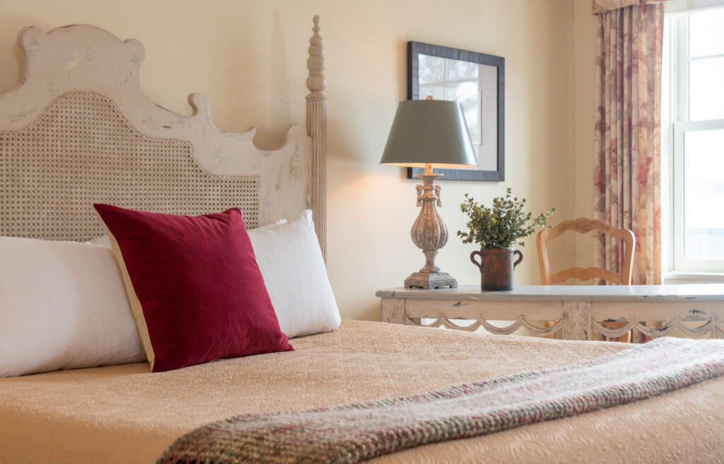 Cozy bedroom with a beige bed, burgundy pillow, lamp, potted plant, wooden desk, and a window letting in natural light.