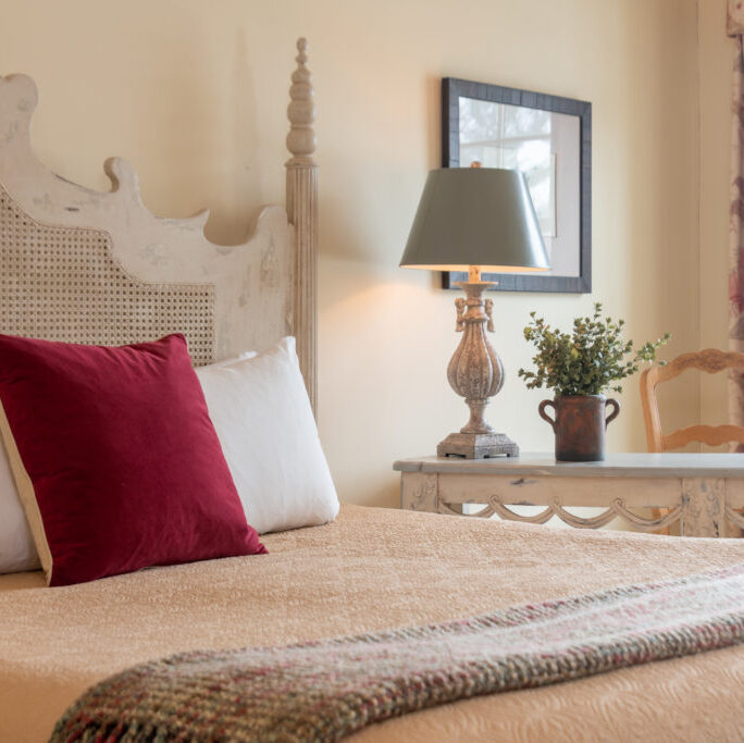 Cozy bedroom with a beige bed, burgundy pillow, lamp, potted plant, wooden desk, and a window letting in natural light.