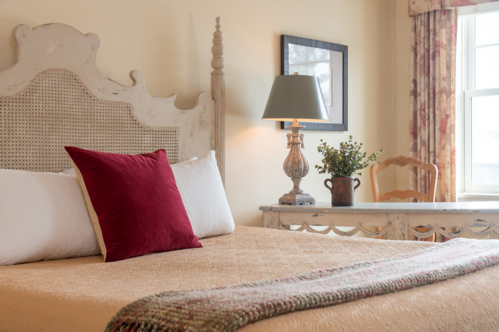 Cozy bedroom with a beige bed, burgundy pillow, lamp, potted plant, wooden desk, and a window letting in natural light.