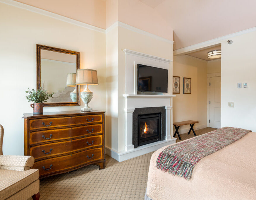 Cozy bedroom with a fireplace, TV, wooden dresser, armchair, bed with a plaid throw, and beige walls with framed artwork and a mirror.