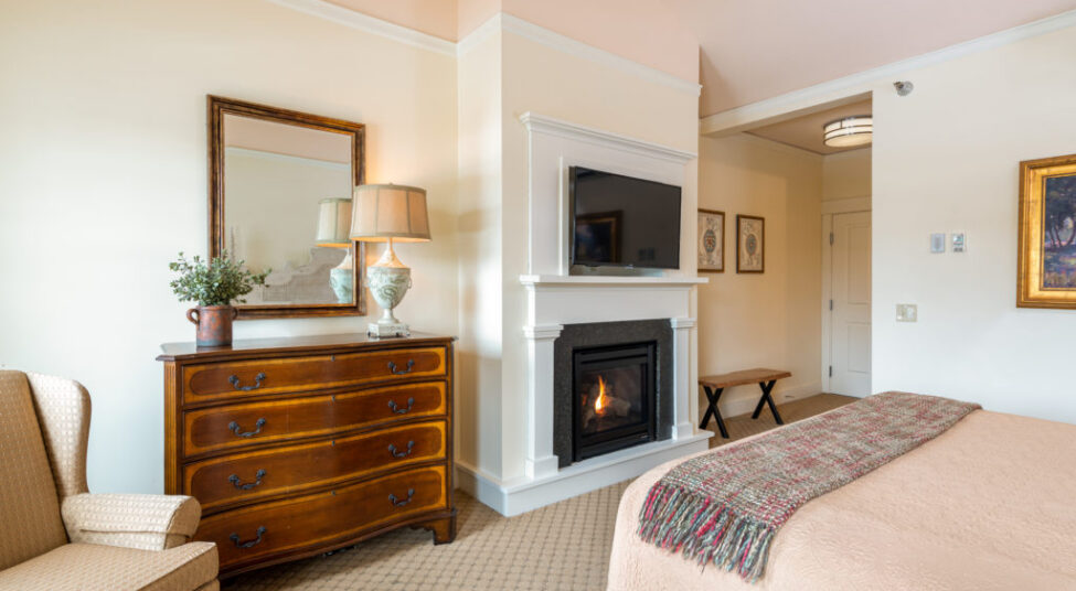 A cozy bedroom with a bed, fireplace, TV, wooden dresser with a lamp and mirror, armchair, and artwork on the walls.