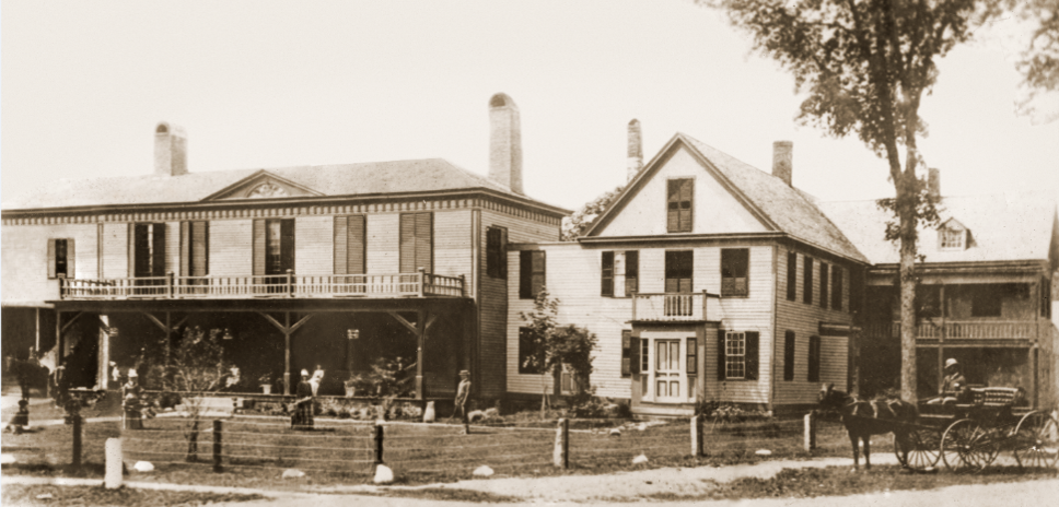 Sepia-toned photo of a large two-story wooden building with balconies, people, and a horse-drawn carriage in front.